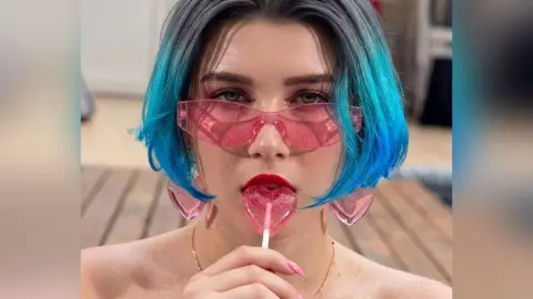 Young woman with blue hair and pink sunglasses biting a heart-shaped lollipop in a bright, playful mood.