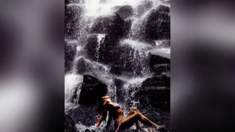 Aalyahh poses naked under a rushing waterfall, showing her wet skin and dark hair while sitting against dark rocks.