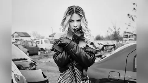 ChristineDavis poses outdoors in a black leather jacket and fishnet top, looking directly at the camera with dark lipstick and wavy blonde hair.