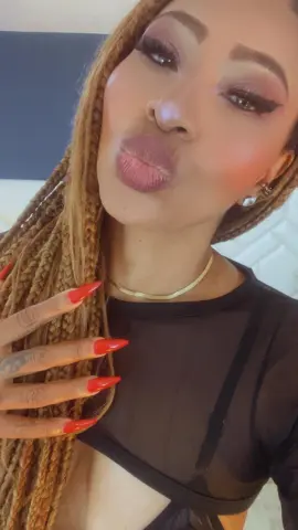 Woman with long blonde braids and red nails wearing a black mesh top making a playful kissy face.