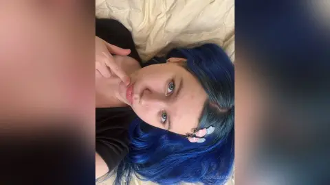 DeidreSarin lying on a bed with her blue hair spread out, touching her chin and looking at the camera.