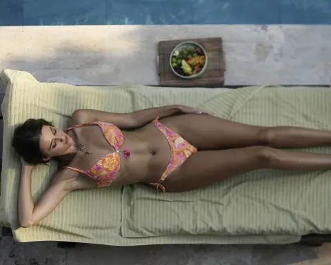 Relaxed woman in a colorful patterned bikini lounging on a sunbed by a poolside with fresh fruit.