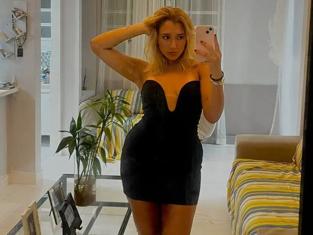 Blonde woman in a chic black mini dress taking a mirror selfie in a bright, modern living room.