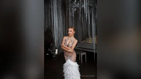 StellaFletcher poses in a white, feathered, low-cut gown against a dark background with hanging white tinsel or fringe.
