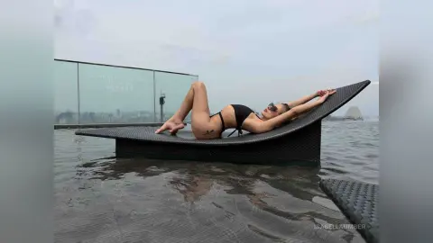 Relaxed woman in black bikini lounging on a sunbed at a rooftop infinity pool overlooking a city skyline.