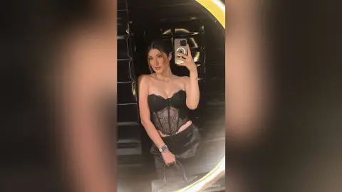 Young woman in a black lace corset top and dark trousers taking a mirror selfie in a modern dark interior.