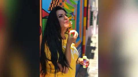 MissyJolie smiles while holding a tube of lip gloss near her mouth, wearing a bright yellow t-shirt with long black hair.