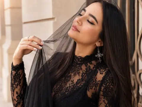 Elegant woman in a black lace dress and veil posing with a serene expression against a stone building.
