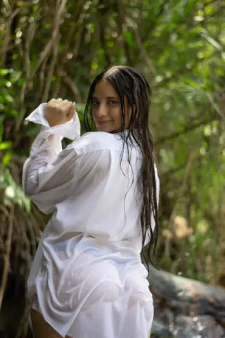 Smiling woman in a white linen shirt posing in a lush green tropical forest setting.