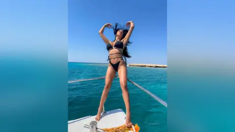 NicoleBella stands on a boat deck in a black bikini against a clear blue ocean and sky.