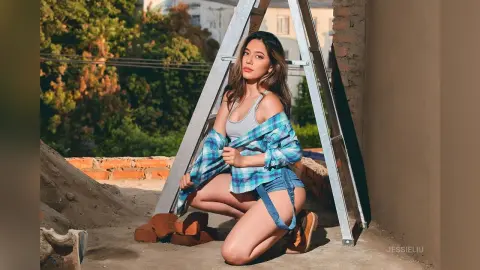 JessieLiu kneels on a rooftop wearing a grey tank top and blue plaid shirt left open to reveal her chest, paired with denim shorts.