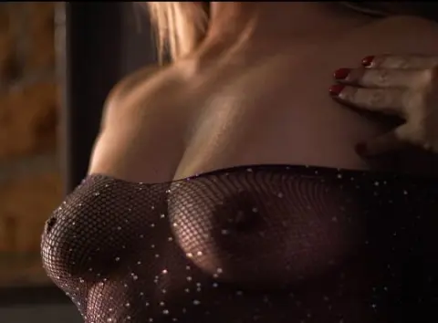 Woman in sheer sparkly mesh top posing in warm, moody indoor lighting.
