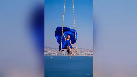 Woman in a flowing blue dress swinging high over the ocean against a clear blue sky.