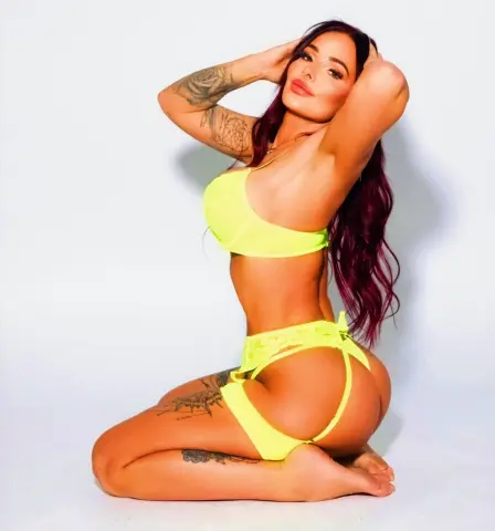 SOAKED_GYMGIRL_MILF_UK poses in neon yellow lingerie, showing off her tattoos and long red hair while kneeling against a white background.