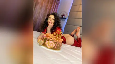 Glamorous woman in red silk pajamas celebrating her birthday with a pink cake on a white bed.