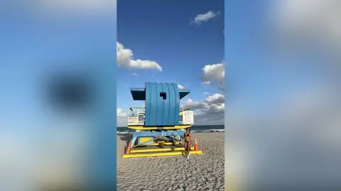 Woman in a black bikini relaxing on a sunny Miami Beach sand near a blue lifeguard tower under a clear sky.