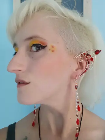 Woman with blonde hair wearing red gemstone statement earrings and elven ear cuffs against a blue background.