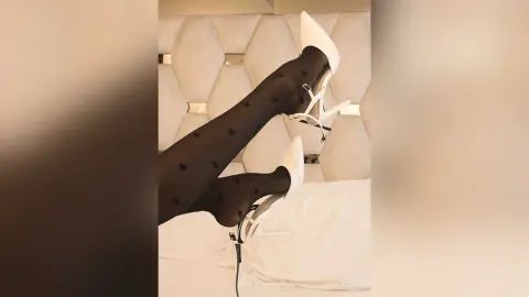 Woman in black patterned tights and white high heel pumps relaxing on a white bed in a bright room.