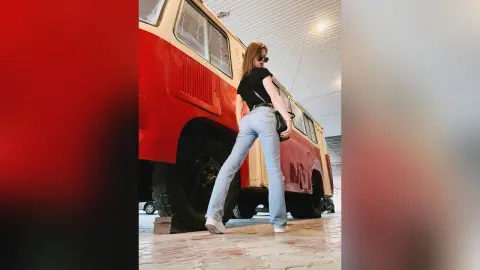Stylish woman in black top and light blue jeans posing next to a vintage red bus in a bright garage.