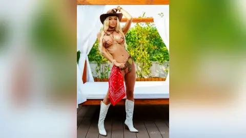 Anastasia stands outdoors wearing a brown cowboy hat, a brown bikini top, and a red bandana tied around her waist with white cowboy boots.