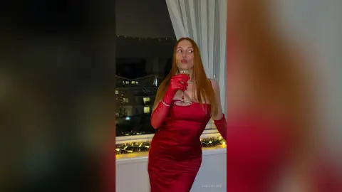 Elegant redhead in a red satin evening gown holding a champagne glass in a festive nighttime setting.