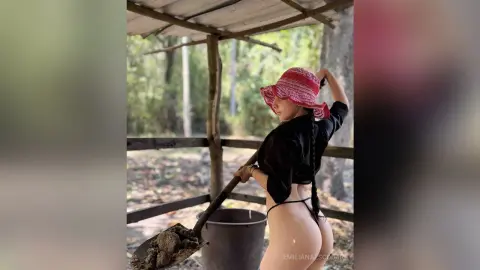 Young woman in a black top and red sun hat relaxing under a rustic wooden shelter in a lush forest setting.