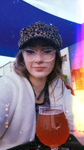 jenellehayesxoxo wears a leopard print cap and clear glasses while posing with a glass of red liquid.