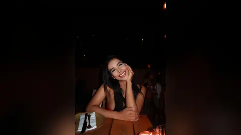 Alison smiles while sitting at a dark restaurant table, resting her chin on her hand with her long dark hair down.