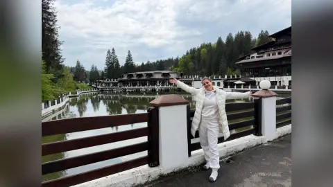 AnishaBee stands outside with her arms spread wide, wearing a white puffer jacket and white pants against a backdrop of a lake and forest.