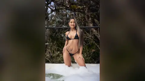 Smiling woman in a black bikini relaxing in a white hot tub surrounded by lush green forest trees.