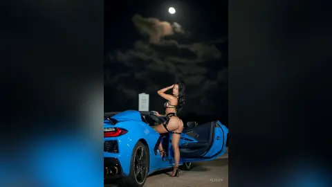 Alison poses next to a blue convertible car at night, wearing a black strappy lingerie set that shows her bare back and buttocks.