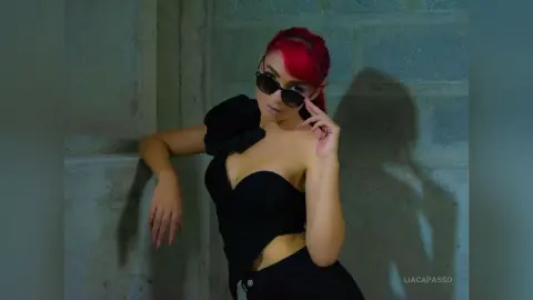 Red-haired woman in a black strapless top posing against a concrete wall in moody lighting.