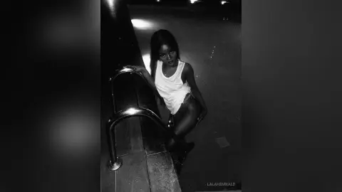 Stylish woman in a white tank top posing on stairs in a moody, dark cinematic setting.