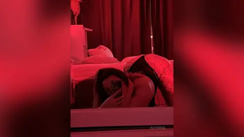 Woman in black fishnet lingerie relaxing on a bed in a moody, red-lit bedroom setting.