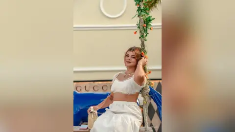 Smiling woman in a white crop top and skirt sitting on a floral swing in a bright, elegant indoor setting.