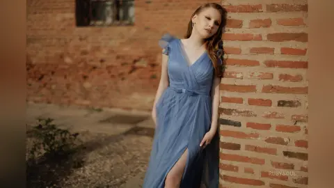RubySoul poses leaning against a brick wall wearing a long, light blue dress with a high slit and ruffles.
