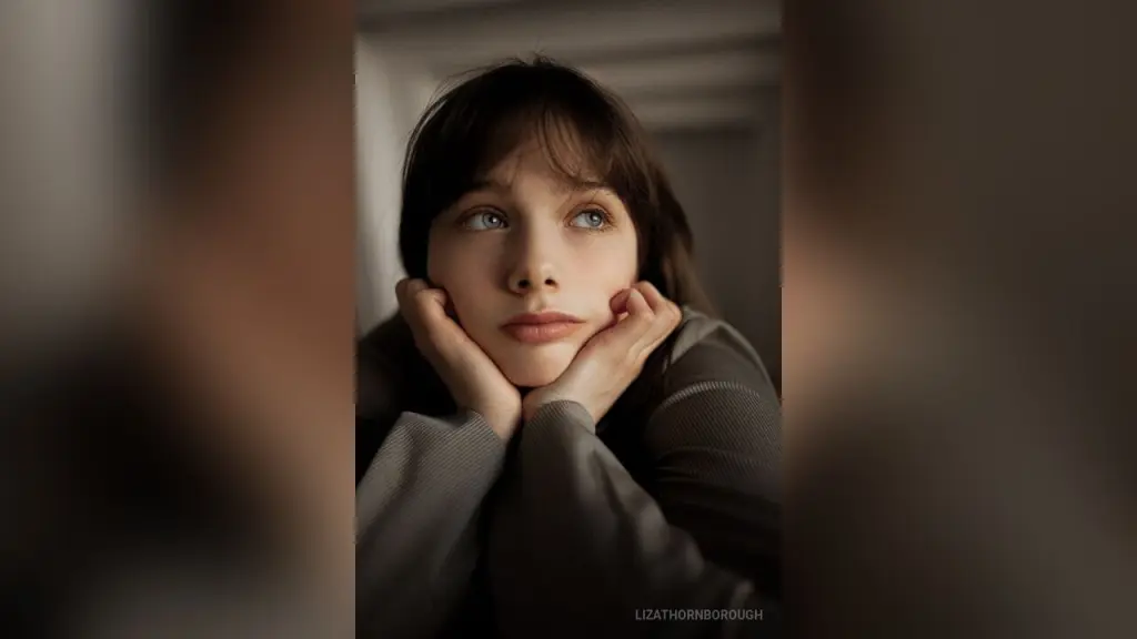 Pensive young woman with brown hair and blue eyes resting her chin on her hands in a moody indoor setting.