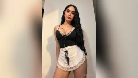Young woman with long wavy hair wearing a black lace camisole and white ruffled apron in a minimalist setting.