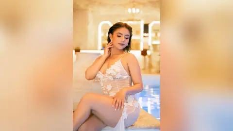 Elegant woman in a white floral lace dress posing gracefully in a luxurious, sunlit indoor setting.