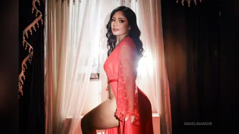 Elegant woman in a flowing red sheer robe posing by a window in soft, romantic lighting.
