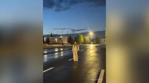 MagalyBoffa stands on a wet, dark road at dusk, wearing light-colored pants and a long-sleeved top with her long hair down.