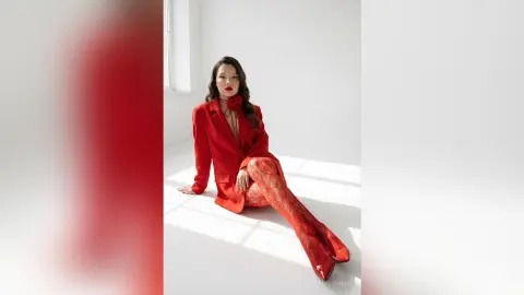 Elegant woman in a red blazer and patterned tights sitting in a bright, minimalist white studio.