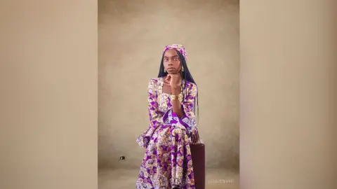Elegant Black woman in a purple patterned maxi dress posing thoughtfully against a neutral studio background.