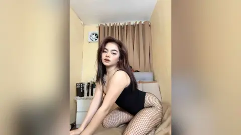 Young woman in a black bodysuit and fishnet tights posing in a cozy bedroom setting.