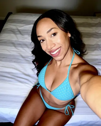 MistressJacksonMonroe smiling in a blue polka dot bikini while sitting on a striped bed.