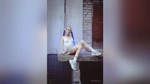 Young woman in white tank top and plaid skirt sitting on a concrete ledge in an urban alleyway.