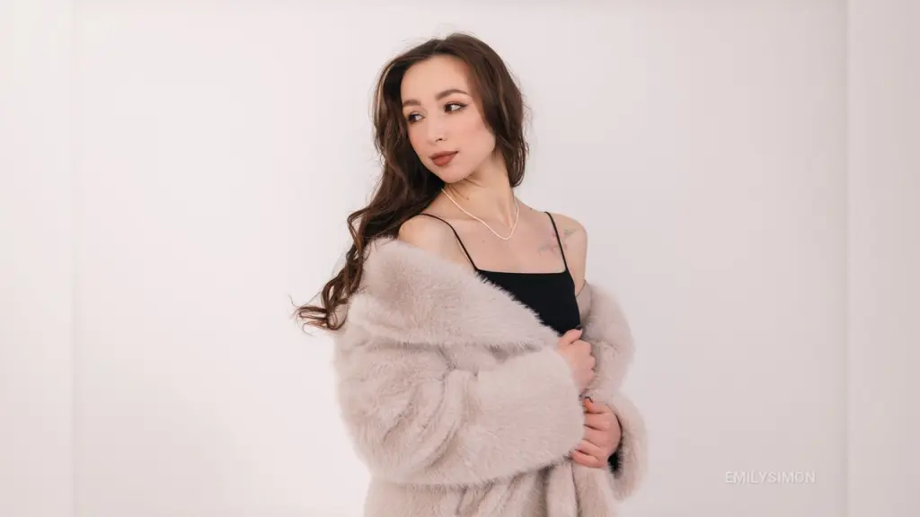 EmilySimon — Elegant woman in a black camisole and plush beige faux fur coat posing against a white studio background.