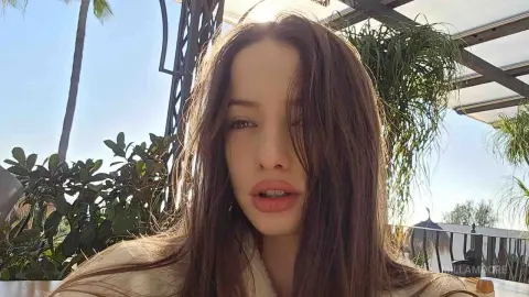 Young woman with long brown hair in a casual beige outfit relaxing outdoors in a sunny tropical garden setting.