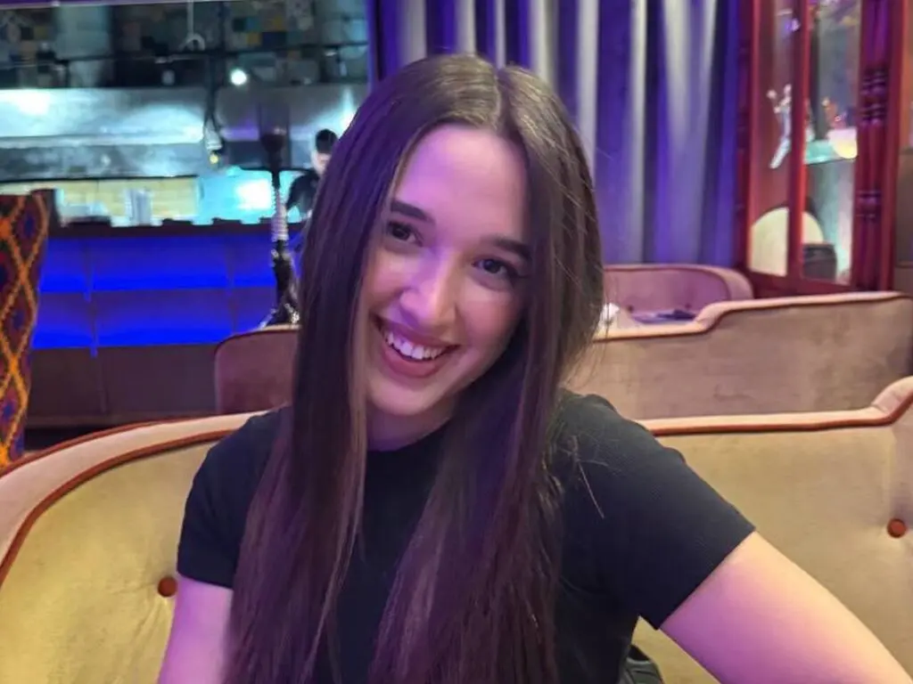 MaryamKeets smiles while wearing a black t-shirt, showing her long brown hair in a room with soft lighting and a tan couch.