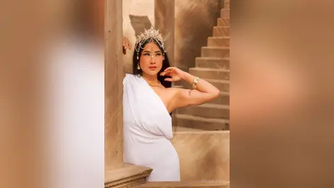 JuliahLewis wears a white one-shoulder dress and a gold tiara while posing against a stone wall and stairs.